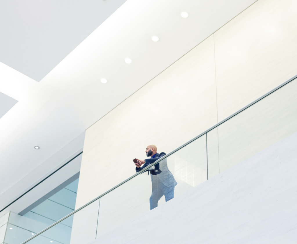 A man in a suit stands on an upper-level balcony in a modern, brightly lit building, checking his phone and reviewing financial checklists.